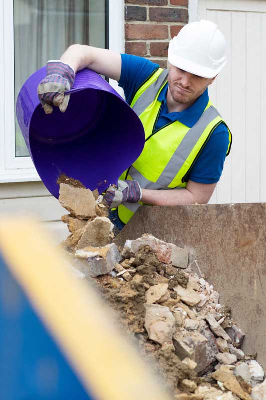Construction worker wearing a hard-hat emptying building waste into skip outside renovated house