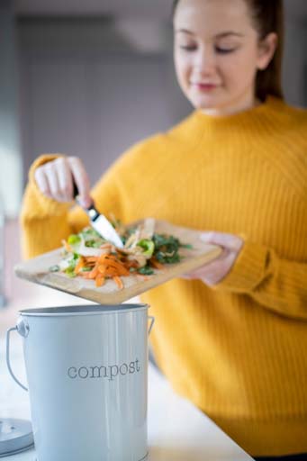 Woman in kitchen placing vegetable leftovers from a cutting board into a composting pail